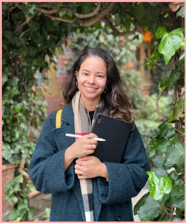 Woman holding a black folder and pen outdoors with greenery in the background